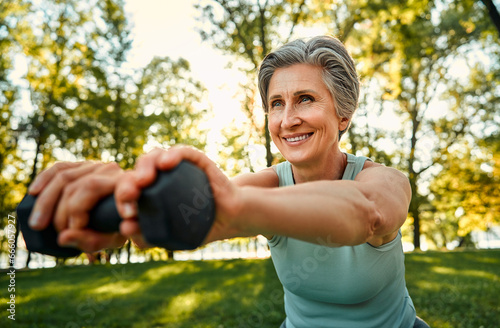 Regular workout outdoors. Close up of smiling aged lady with stylish haircut training on fresh air with dumbbells. Sporty woman strengthening muscles and bones with slight exercises.