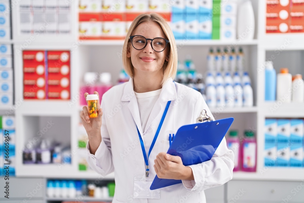 Young caucasian woman working at pharmacy drugstore holding pills smiling looking to the side and staring away thinking.