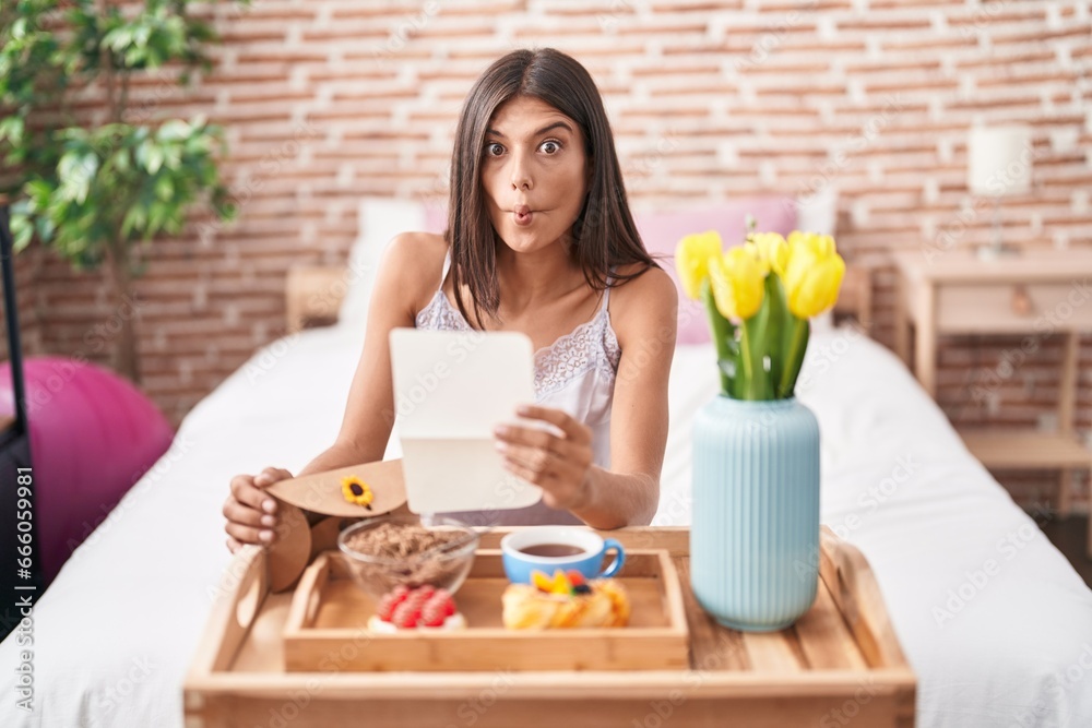 Brunette young woman eating breakfast in the bed reading a letter making fish face with mouth and squinting eyes, crazy and comical. Brunette young woman eating breakfast in the bed reading a letter making fish face with mouth and squinting eyes, crazy and comical.
