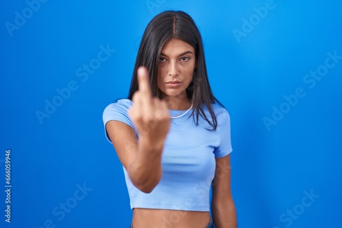 Brunette young woman standing over blue background showing middle finger  impolite and rude fuck off expression