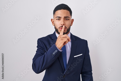 Young hispanic man wearing business suit and tie asking to be quiet with finger on lips. silence and secret concept.