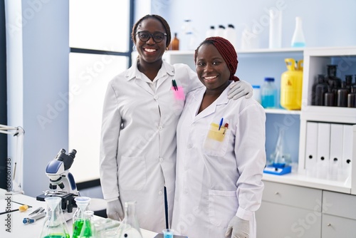 Two african women working at scientist laboratory looking positive and happy standing and smiling with a confident smile showing teeth
