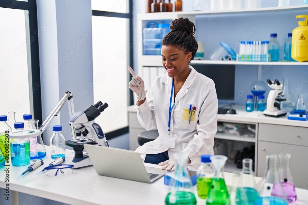 Young african american with braids working at scientist laboratory with laptop smiling happy pointing with hand and finger to the side