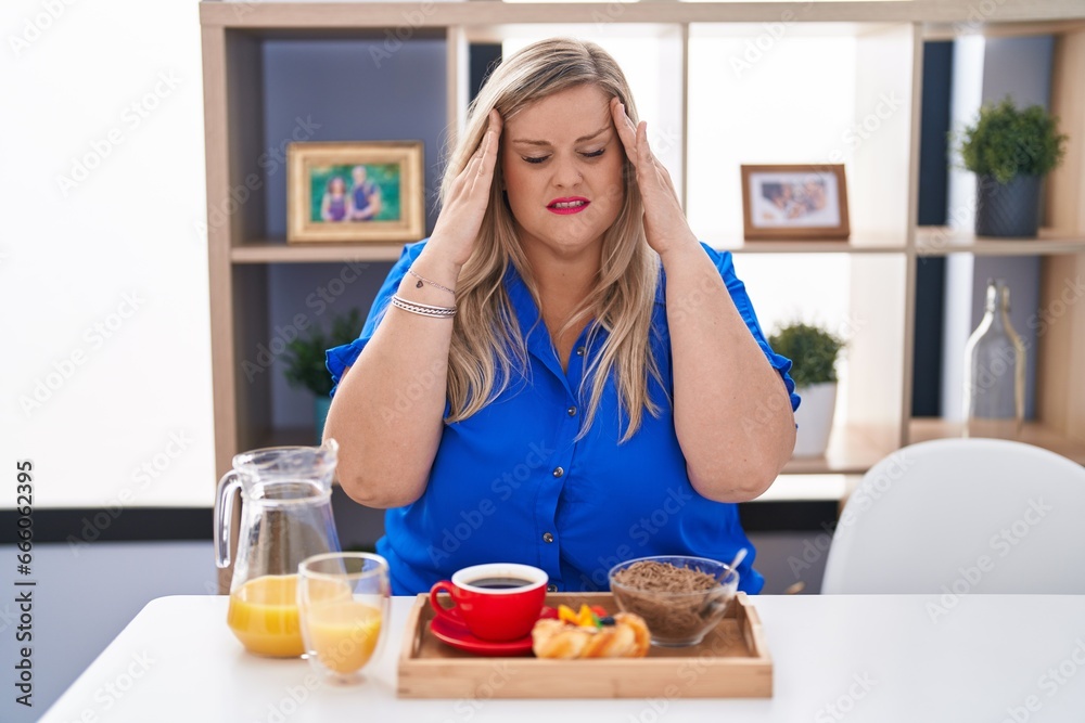 Caucasian plus size woman eating breakfast at home with hand on head for pain in head because stress. suffering migraine. Caucasian plus size woman eating breakfast at home with hand on head for pain in head because stress. suffering migraine.