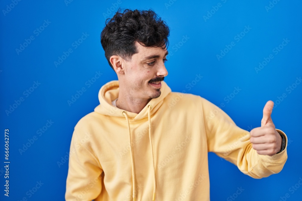 Hispanic man standing over blue background looking proud, smiling doing thumbs up gesture to the side