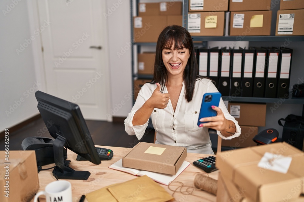 Young brunette woman working at small business ecommerce using smartphone smiling happy and positive, thumb up doing excellent and approval sign Young brunette woman working at small business ecommerce using smartphone smiling happy and positive, thumb up doing excellent and approval sign