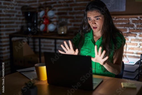 Young teenager girl working at the office at night afraid and terrified with fear expression stop gesture with hands, shouting in shock. panic concept.