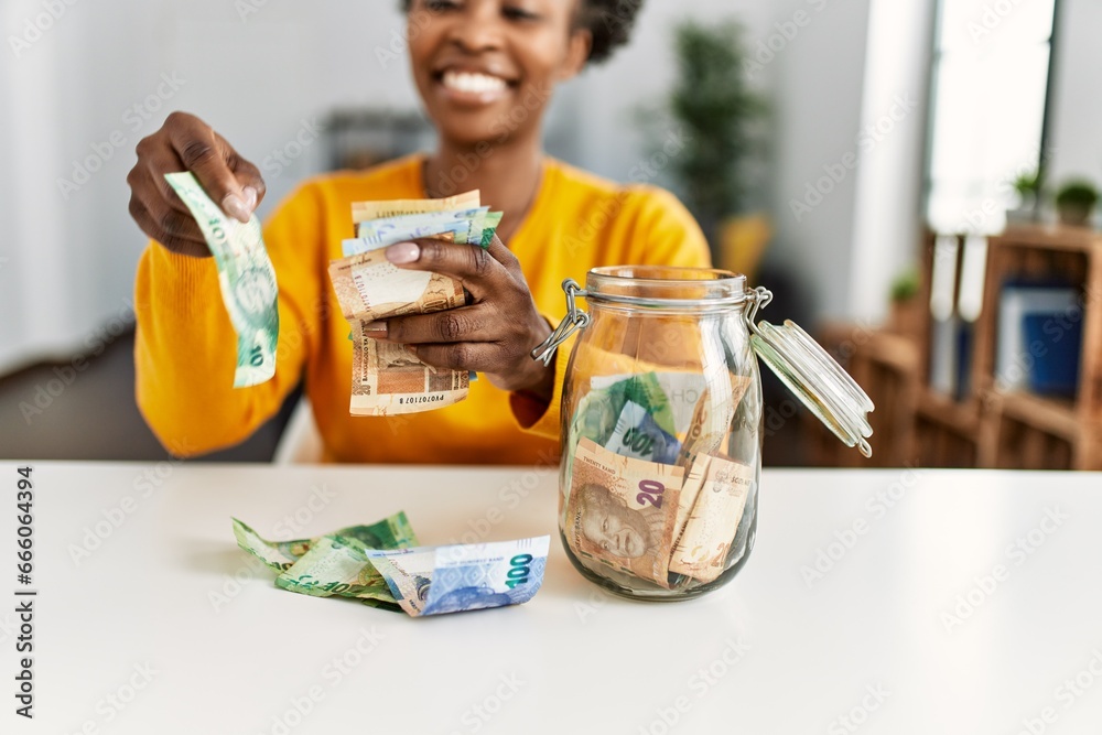 Φωτογραφία African american woman sitting on table holding rand banknotes of charity jar at