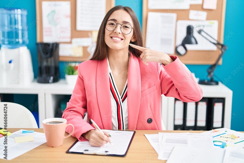 Young hispanic woman working at the office wearing glasses smiling cheerful showing and pointing with fingers teeth and mouth. dental health concept.