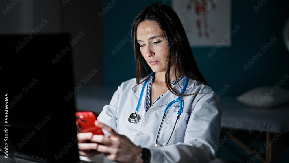 Young beautiful hispanic woman doctor using smartphone working at clinic Young beautiful hispanic woman doctor using smartphone working at clinic