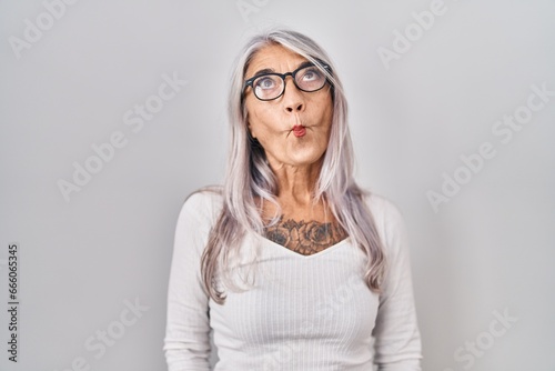 Middle age woman with grey hair standing over white background making fish face with lips, crazy and comical gesture. funny expression.