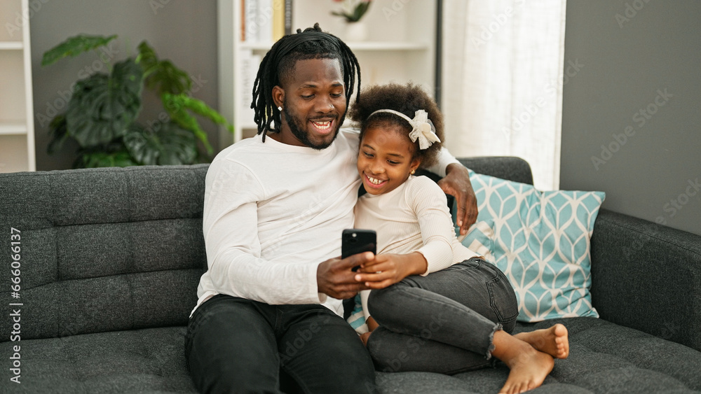 Obraz premium African american father and daughter using smartphone sitting on sofa smiling at home