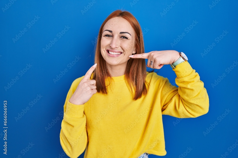 Young woman standing over blue background smiling cheerful showing and pointing with fingers teeth and mouth. dental health concept.