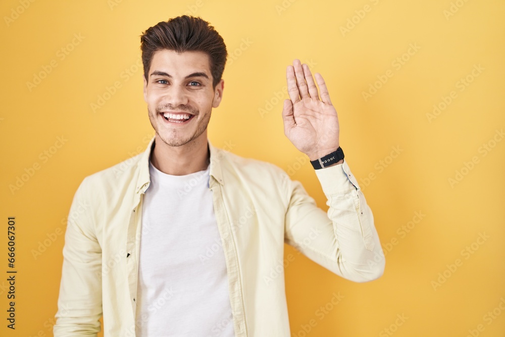 Young hispanic man standing over yellow background waiving saying hello happy and smiling, friendly welcome gesture Young hispanic man standing over yellow background waiving saying hello happy and smiling, friendly welcome gesture