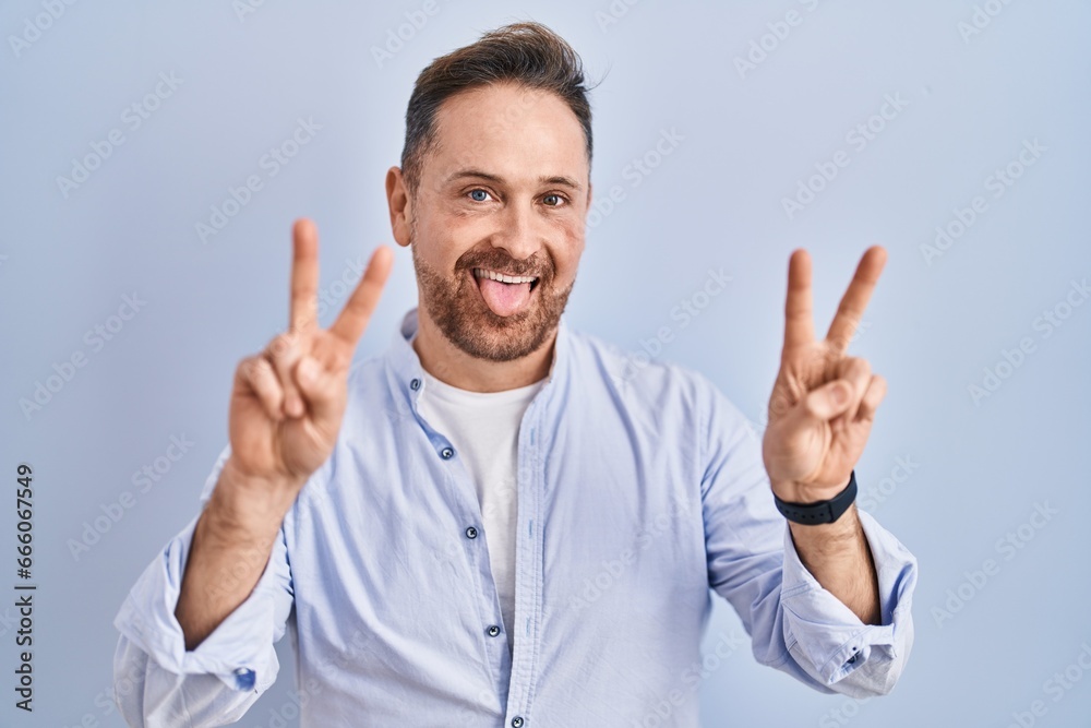 Middle age caucasian man standing over blue background smiling with tongue out showing fingers of both hands doing victory sign. number two. Middle age caucasian man standing over blue background smiling with tongue out showing fingers of both hands doing victory sign. number two.