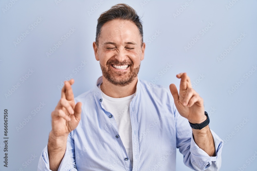 Middle age caucasian man standing over blue background gesturing finger crossed smiling with hope and eyes closed. luck and superstitious concept. Middle age caucasian man standing over blue background gesturing finger crossed smiling with hope and eyes closed. luck and superstitious concept.
