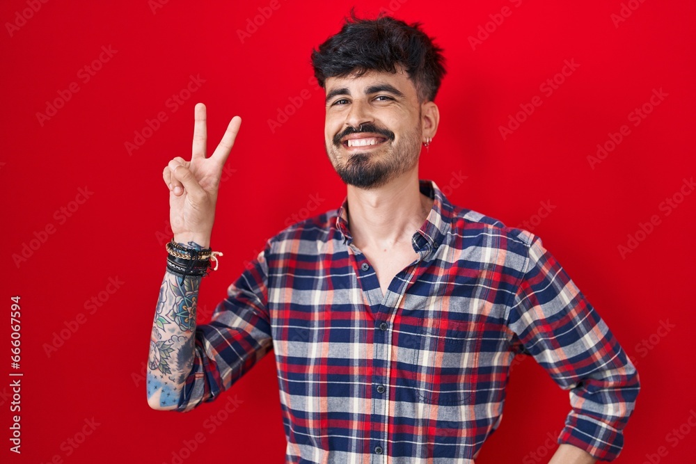 Naklejka premium Young hispanic man with beard standing over red background smiling looking to the camera showing fingers doing victory sign. number two.