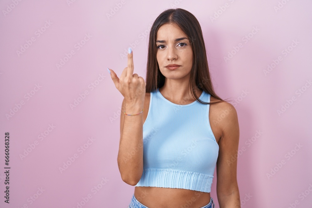 Young brunette woman standing over pink background showing middle finger, impolite and rude fuck off expression