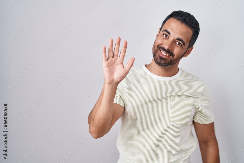 Fototapeta premium Hispanic man with beard standing over isolated background waiving saying hello happy and smiling, friendly welcome gesture