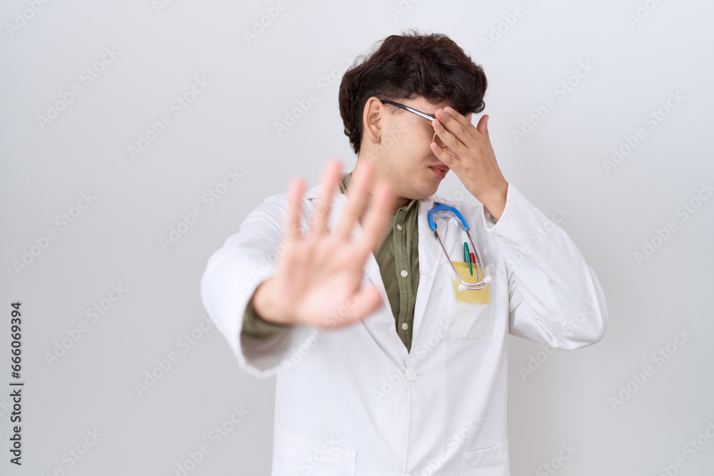 Young non binary man wearing doctor uniform and stethoscope covering eyes with hands and doing stop gesture with sad and fear expression. embarrassed and negative concept. Young non binary man wearing doctor uniform and stethoscope covering eyes with hands and doing stop gesture with sad and fear expression. embarrassed and negative concept.