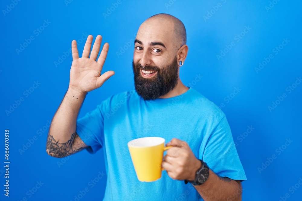 Young hispanic man with beard and tattoos drinking a cup of coffee waiving saying hello happy and smiling, friendly welcome gesture Young hispanic man with beard and tattoos drinking a cup of coffee waiving saying hello happy and smiling, friendly welcome gesture