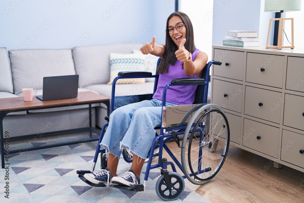 Young hispanic woman sitting on wheelchair at home approving doing positive gesture with hand, thumbs up smiling and happy for success. winner gesture. Young hispanic woman sitting on wheelchair at home approving doing positive gesture with hand, thumbs up smiling and happy for success. winner gesture.