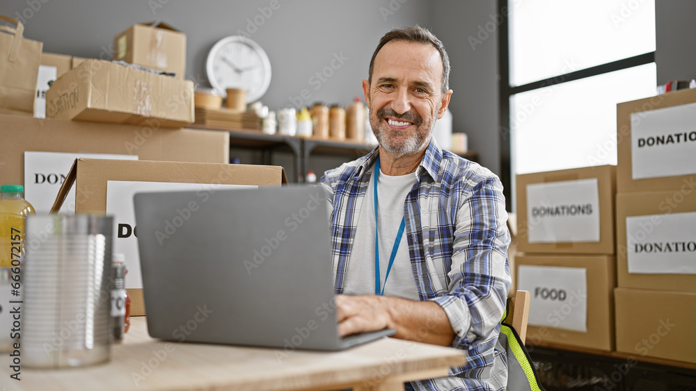 Smiling middle-aged man with grey hair confidently volunteers at a community charity center, exuding unity and altruism while skillfully operating a laptop. Smiling middle-aged man with grey hair confidently volunteers at a community charity center, exuding unity and altruism while skillfully operating a laptop.