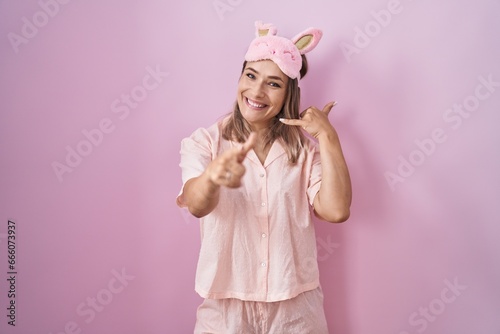 Blonde caucasian woman wearing sleep mask and pajama smiling doing talking on the telephone gesture and pointing to you. call me.