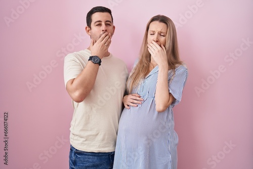 Young couple expecting a baby standing over pink background bored yawning tired covering mouth with hand. restless and sleepiness.