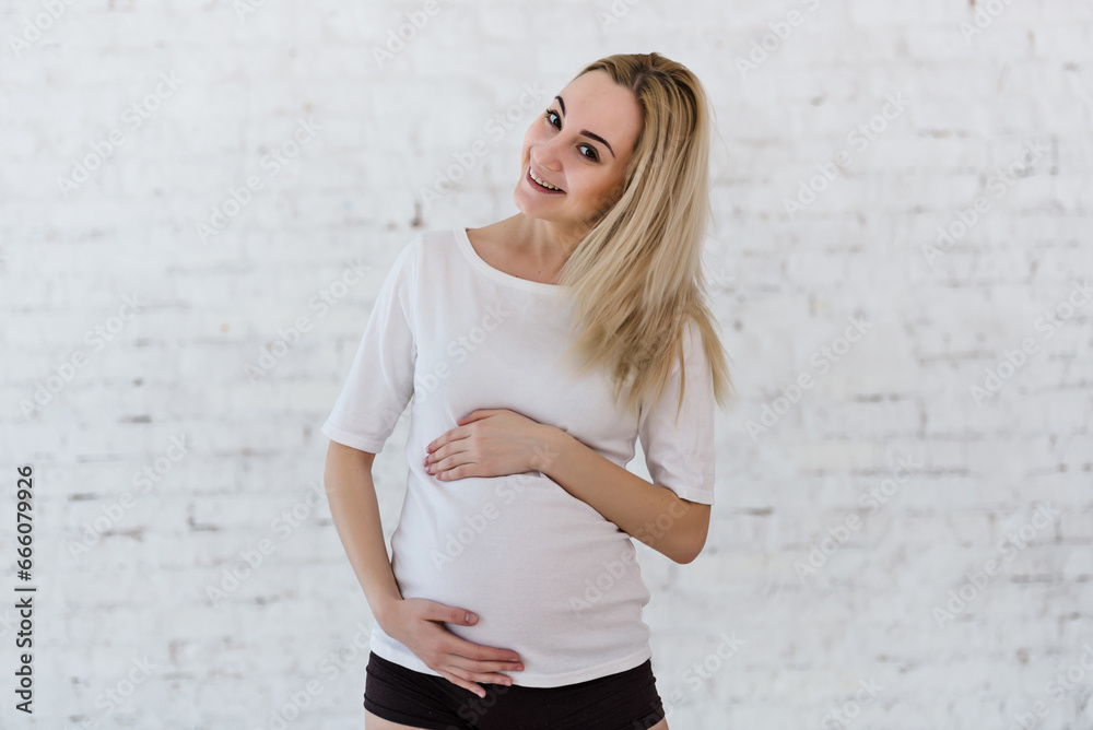 A young blonde pregnant woman stands by the window and strokes her belly in the rays of the morning sun. tender photo session of the expectant mother. pregnant woman in a short top and shorts A young blonde pregnant woman stands by the window and strokes her belly in the rays of the morning sun. tender photo session of the expectant mother. pregnant woman in a short top and shorts