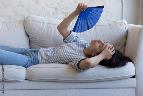 Exhausted sick homeowner woman resting on home sofa, waving handheld paper fan, suffering from fatigue, hypoxia, headache, heating stroke, holding head, swiping sweat