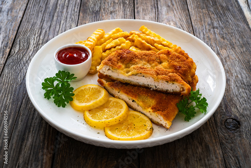 Crispy breaded seared chicken cutlet with French fries, lemon and fresh vegetables on wooden table 