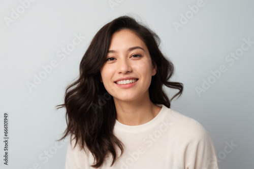 Everyday people. A happy asian woman laughing. Black windswept wavy hair over her shoulder. Wearing a light shirt. Pretty woman. University student. Wholesome. On a grey studio background. Portrait.
