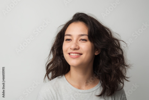 Everyday people. A happy woman smiling. Black wavy hair over her shoulder. Wearing a light shirt. Pretty woman. University student. Wholesome. On a grey studio background. Portrait.