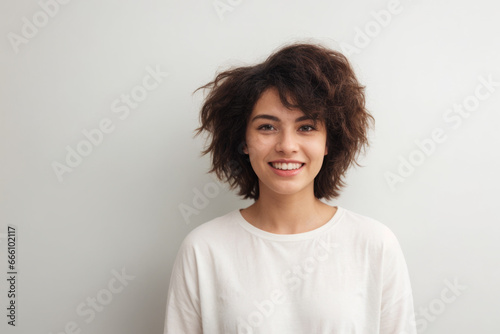 Everyday people. A smiling woman. Brown frizzy hair to her shoulders. Hair volume. Wearing a white shirt. On a light studio background. Portrait.