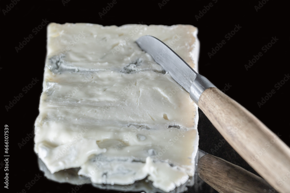 Fromage italien Gorgonzola sur un fond noir avec effet miroir et un couteau à fromage avec un manche en bois posé dessus, différentes couches de bleu et crème apparentes  - obrazy, fototapety, plakaty 