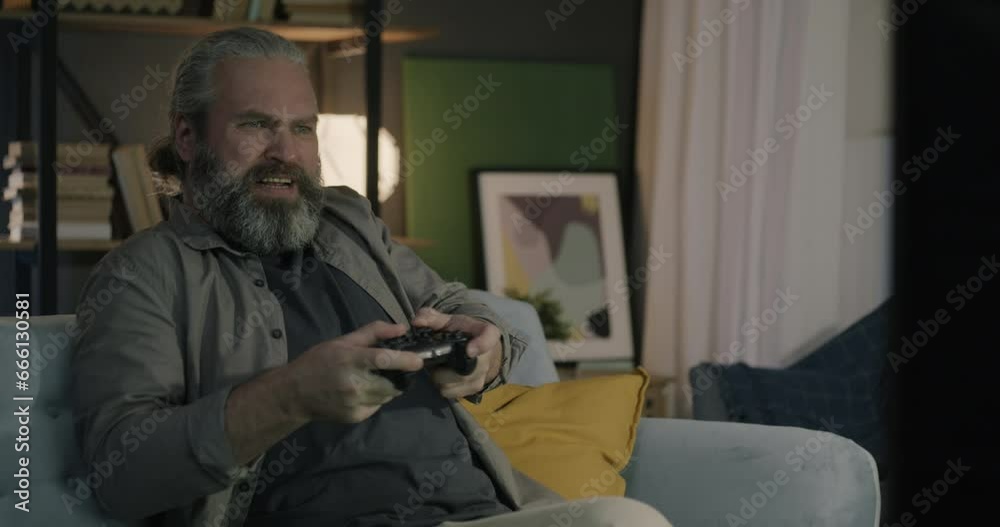 Emotional mature man playing video game with TV console and joystick losing expressing anger sitting on couch in living room at night. People and leisure activity concept. Emotional mature man playing video game with TV console and joystick losing expressing anger sitting on couch in living room at night. People and leisure activity concept.