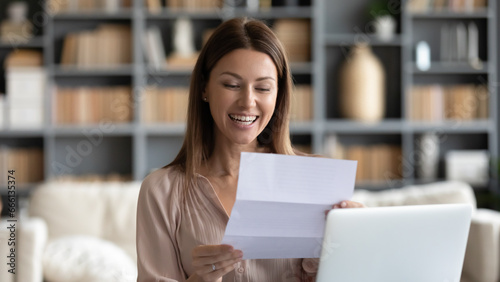Smiling woman reading positive business letter, holding document with good news while sitting at home or modern cozy office desk, housewife got loan approval notification from bank, got job concept