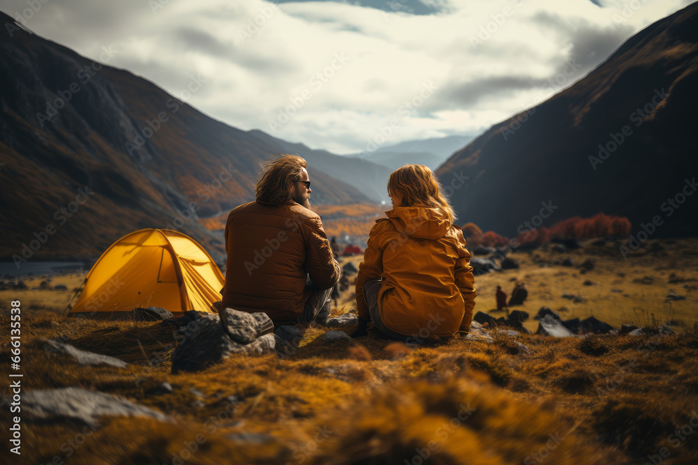 Fototapeta premium Happy couple in outwear sitting on the rocks near the tent camp against the backdrop of picturesque mountain landscape. Caucasian family is engaged in a mountain hiking. Tourism and active lifestyle.
