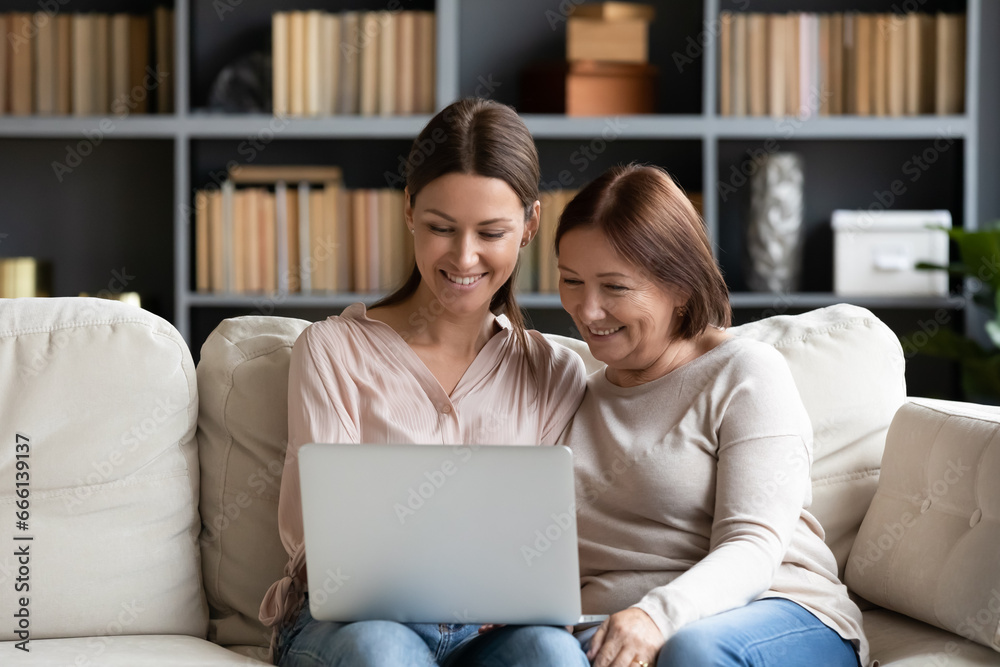 Grown up daughter help aged mum with pc usage, shows her how make video call, multi-generational women family seated on couch buying together on-line, use easy convenient services in internet concept