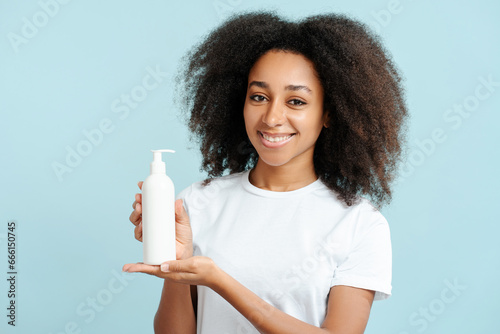 Portrait of happy African American woman with curly hair holding white shampoo bottle, mockup © Maria Vitkovska
