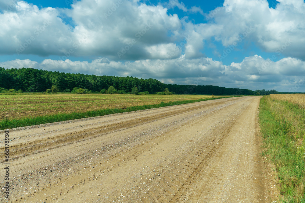 A dry sandy road passes through a field under the scorching sun and clouds. Dirt road outside the city in the village. Arid climate on earth. Climate change and its consequences.