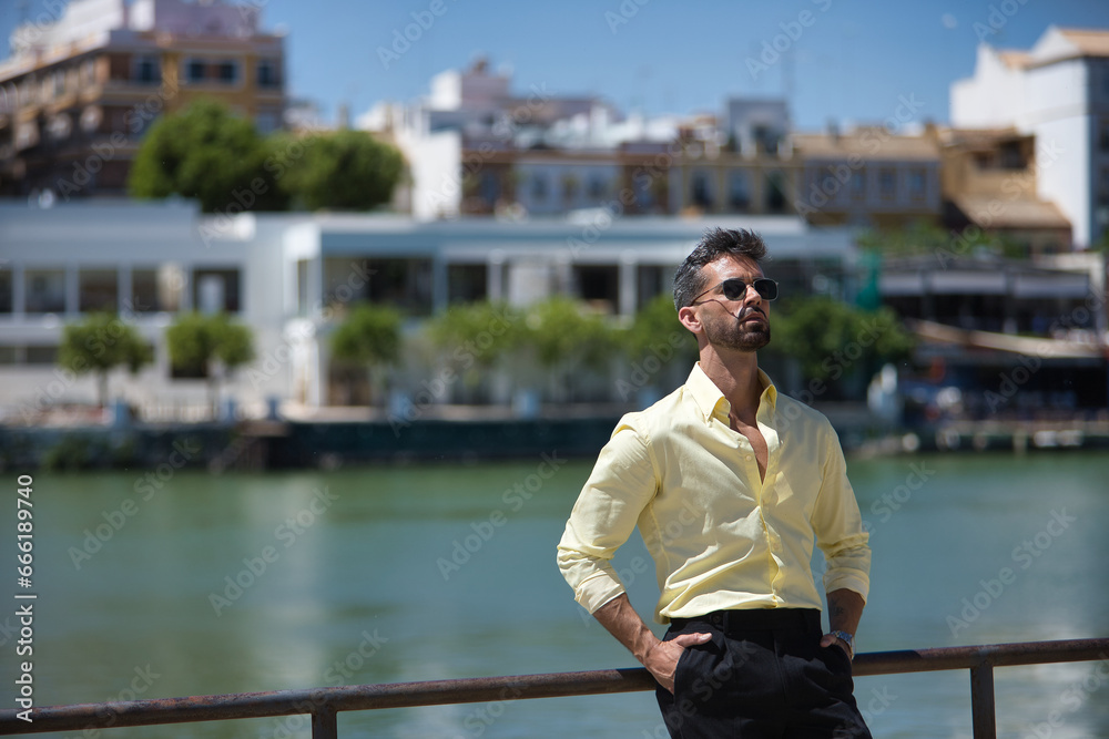 Young and attractive businessman, with beard, yellow shirt, black pants and sunglasses, with his hands in his pockets and looking at the sky leaning on a railing. Concept beauty, fashion, success.