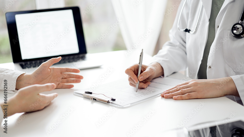 Doctor and patient sitting and discussing something near each other at the white desk in clinic. Female physician is listening filling up a records form. Medicine concept Doctor and patient sitting and discussing something near each other at the white desk in clinic. Female physician is listening filling up a records form. Medicine concept