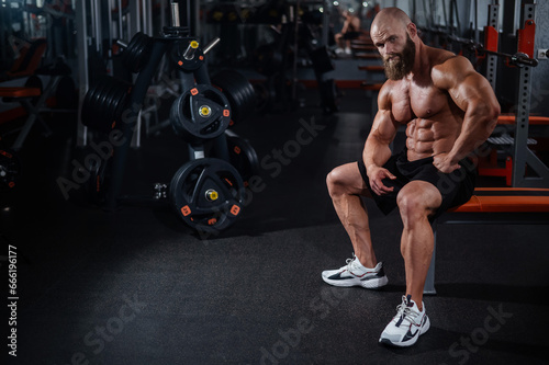 A muscular bald man in shorts is resting on a bench after a workout. Bodybuilder showing off his shape in the gym. 