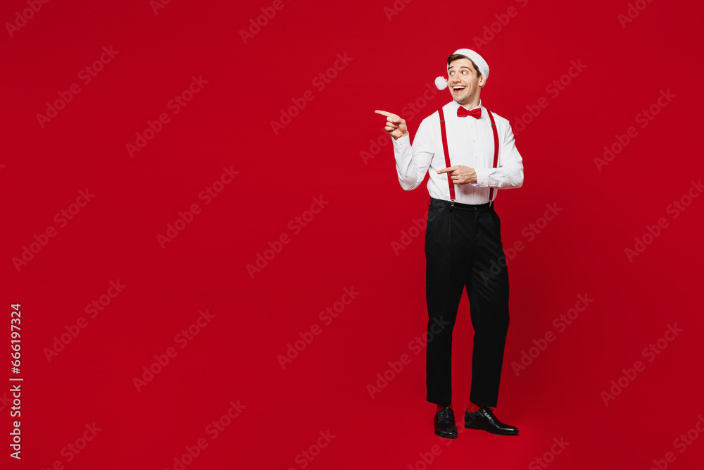 Full body side view merry young man wears white shirt Santa hat posing point index finger aside on area isolated on plain red background studio. Happy New Year Christmas celebration holiday concept. Full body side view merry young man wears white shirt Santa hat posing point index finger aside on area isolated on plain red background studio. Happy New Year Christmas celebration holiday concept.