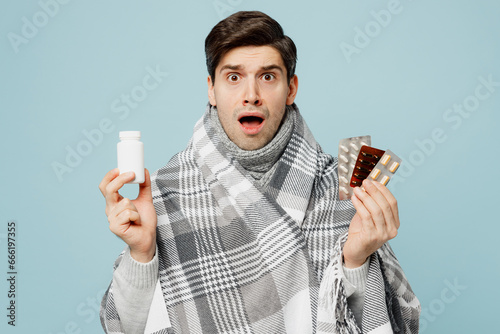 Fototapeta Young scared ill sick man wrapped in gray plaid hold in hand bottle of pills isolated on plain blue background studio portrait