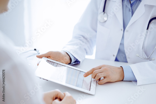 Doctor and patient sitting opposite each other at the desk in clinic. The focus is on female physician s hands pointing into tablet computer touchpad  close up. Medicine concept