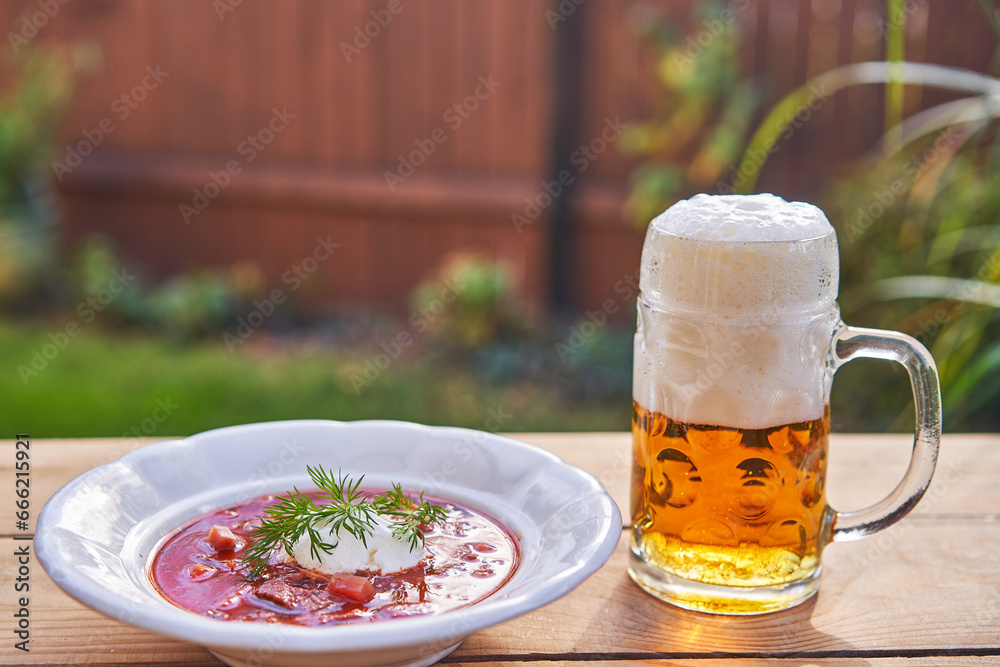 Russian or ukrainian borsch soup served outside with mug of pilsner beer in the garden restaurant in summer sunny day. Beetrot soup with sour cream is traditional recipe from east eauropean cuisine. Russian or ukrainian borsch soup served outside with mug of pilsner beer in the garden restaurant in summer sunny day. Beetrot soup with sour cream is traditional recipe from east eauropean cuisine.