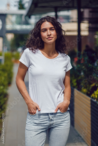 Attractive brunette woman with curly hair in a white t-shirt walks on the street. © kanashkin